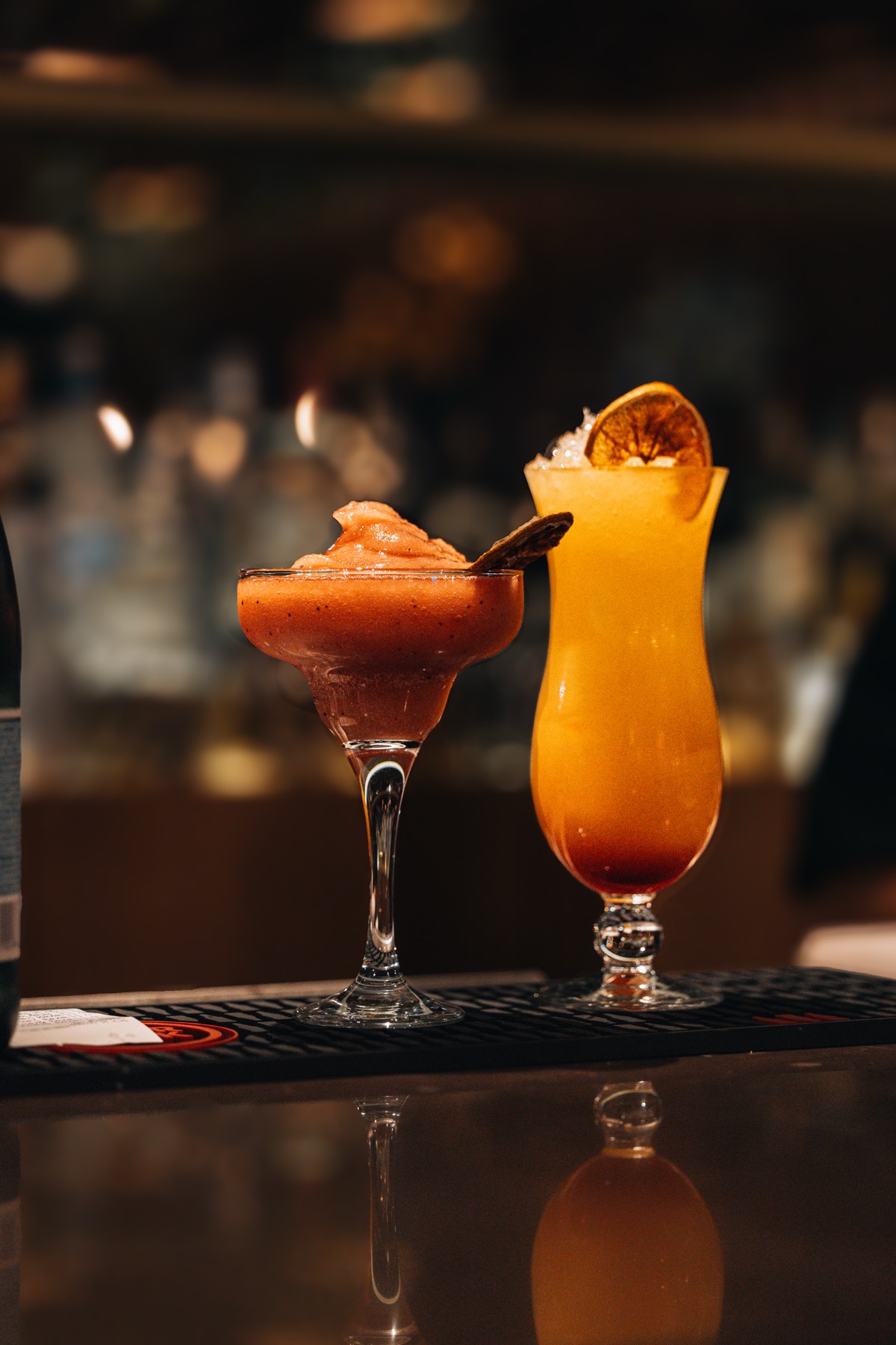 Dois cocktails no balcão do bar, um frozen rosa e um cítrico alaranjado