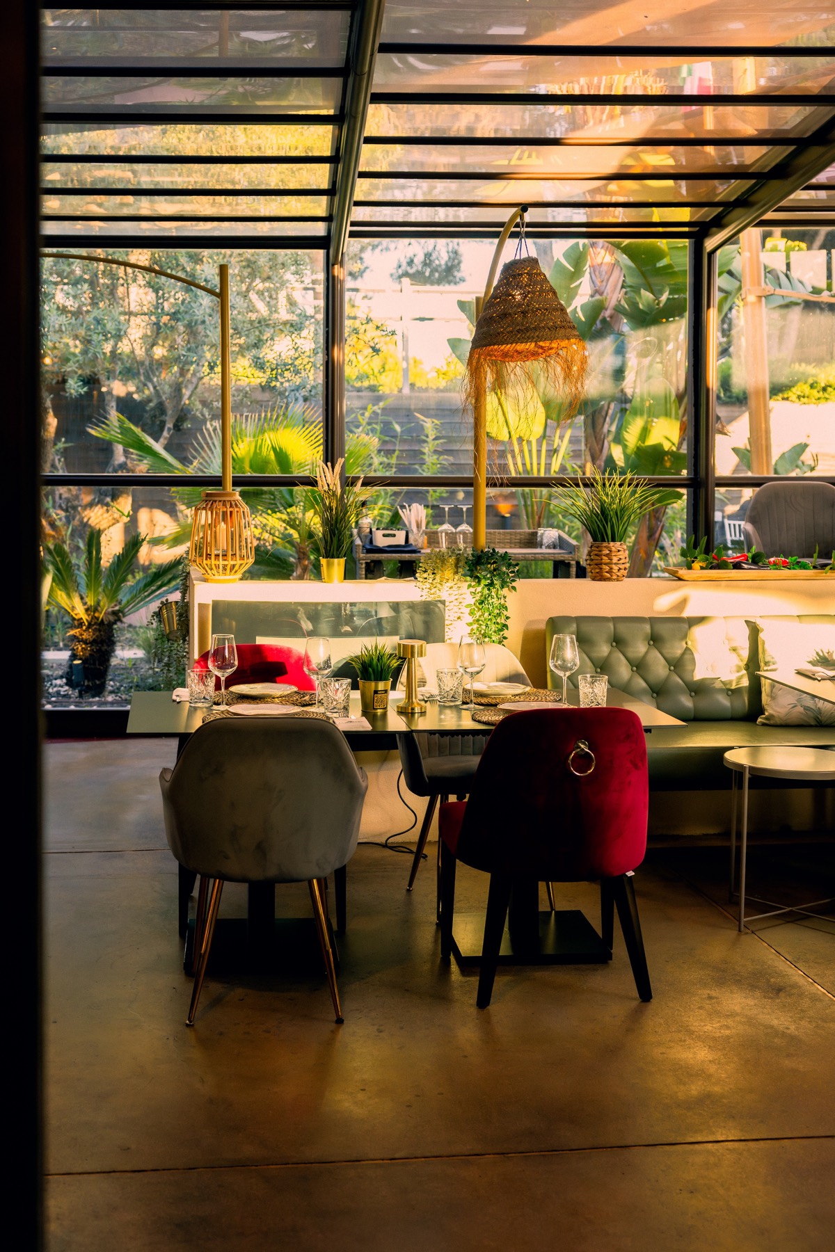 Sala envidraçada do La Bella Fiore com mesa posta, cadeiras de veludo vermelho e cinzento e candeeiro de palha, rodeada de vegetação