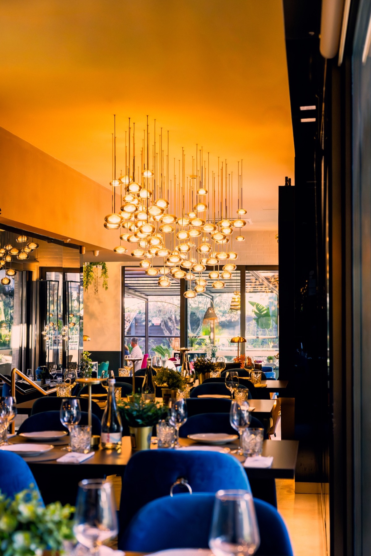 Sala de jantar do La Bella Fiore ao entardecer, com o grande candeeiro em cascata de esferas douradas sobre as mesas postas e cadeiras de veludo azul